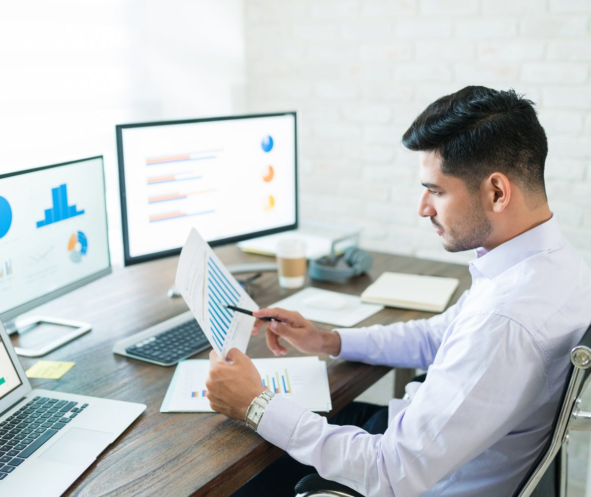 A data analyst reviewing printed charts at a desk with dashboards on screen